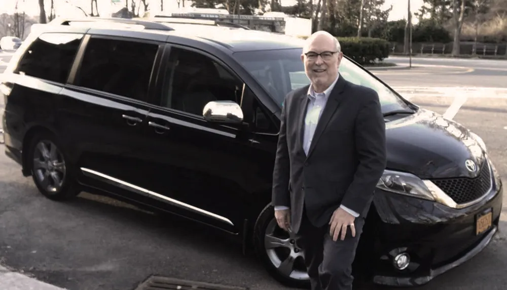 A smiling man in business attire is standing next to a black minivan in a parking area