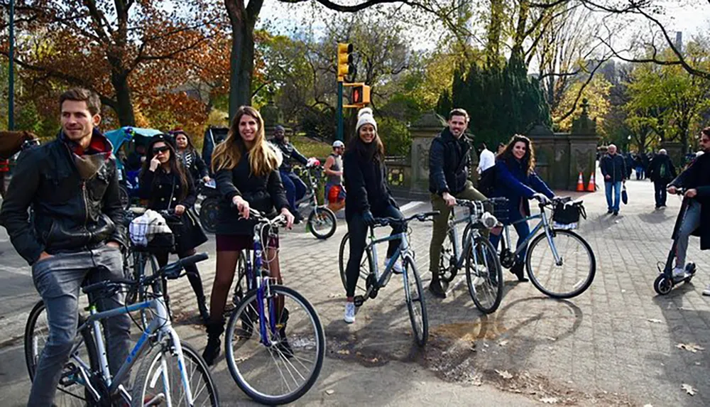 A group of people are smiling and posing on their bicycles at a park with pedestrians in the background