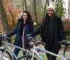 Two smiling women are standing with their bicycles in a park with autumn foliage in the background
