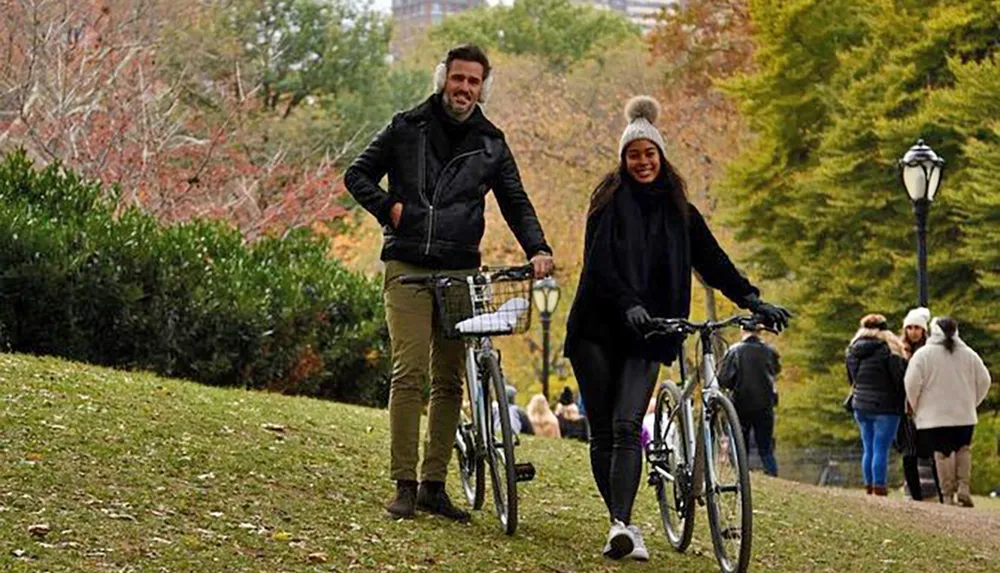 A smiling man and woman walk side by side each pushing a bicycle in a park adorned with autumn foliage