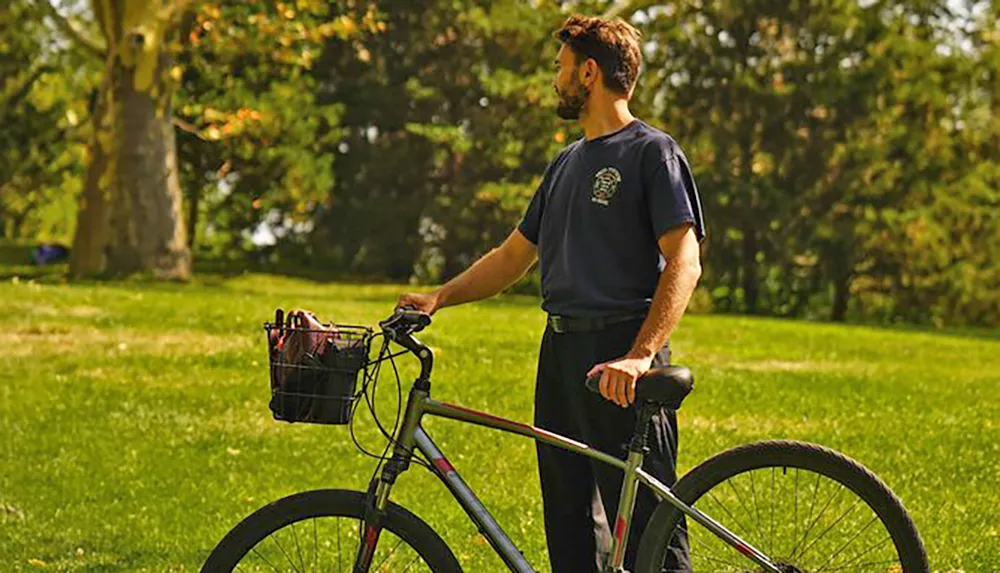 A man stands beside his bicycle in a lush park appearing contemplative or enjoying the scenery