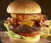 The image shows a cheeseburger with bacon lettuce and tomato accompanied by fries in the background presented on a dark backdrop