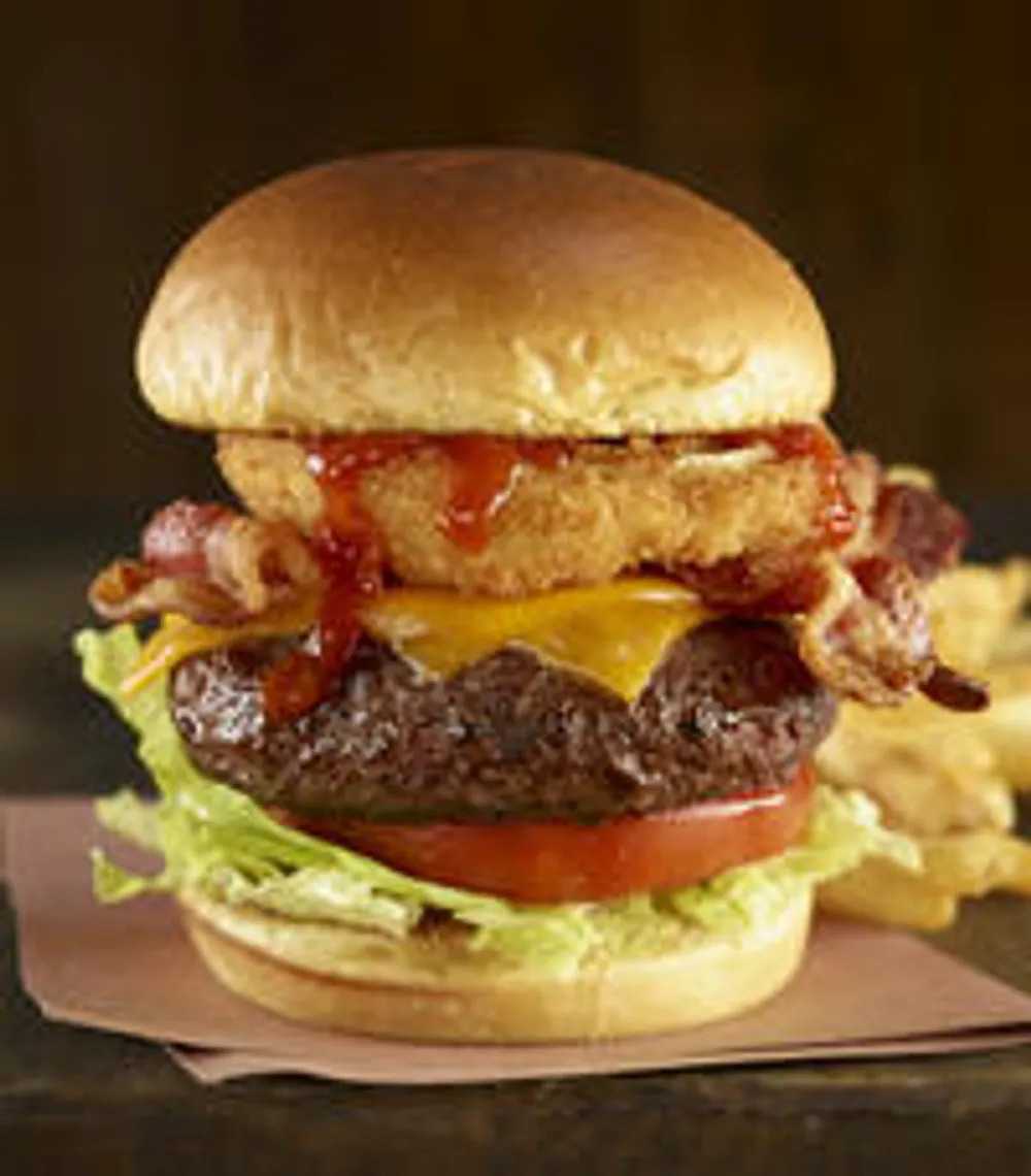 The image shows a cheeseburger with bacon lettuce and tomato accompanied by fries in the background presented on a dark backdrop