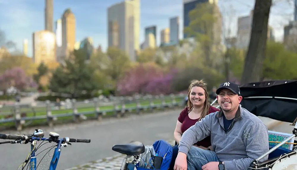 A man and a woman are smiling while sitting next to each other with a bicycle in the foreground and a blurred city park landscape in the background