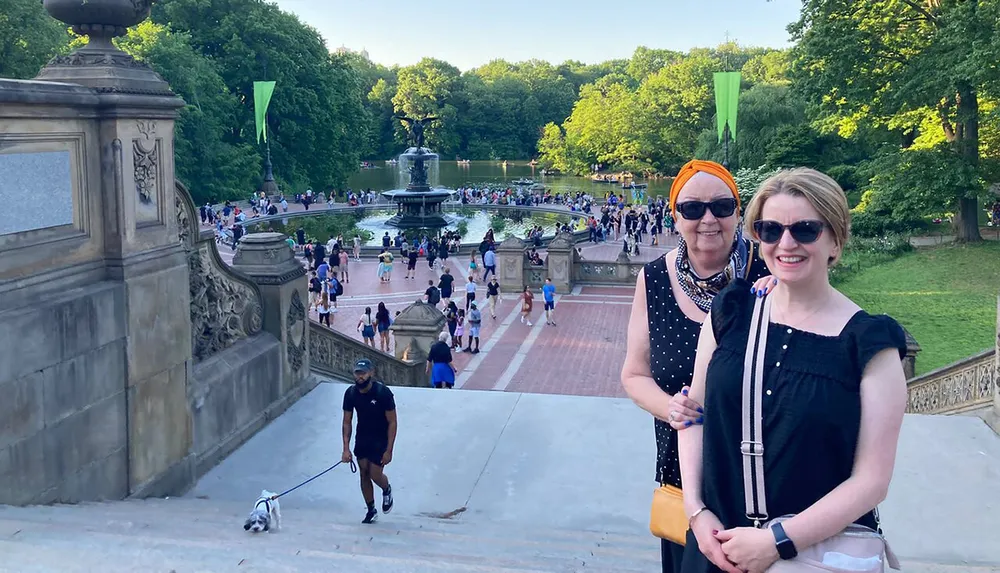 Two people are posing for a photo on a sunny day at a bustling park with steps and a fountain in the background