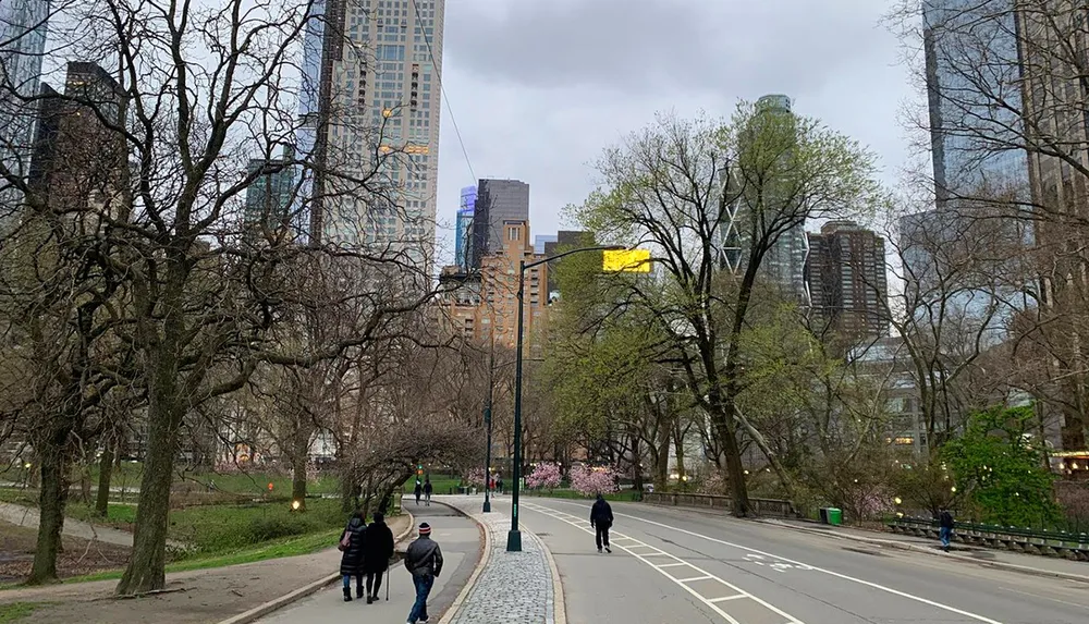 The image captures a tranquil urban park scene with pedestrians walking along a path bare trees early signs of spring and the contrasting backdrop of towering skyscrapers