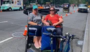 Two people are smiling for a photo while sitting in a pedicab on a city street.