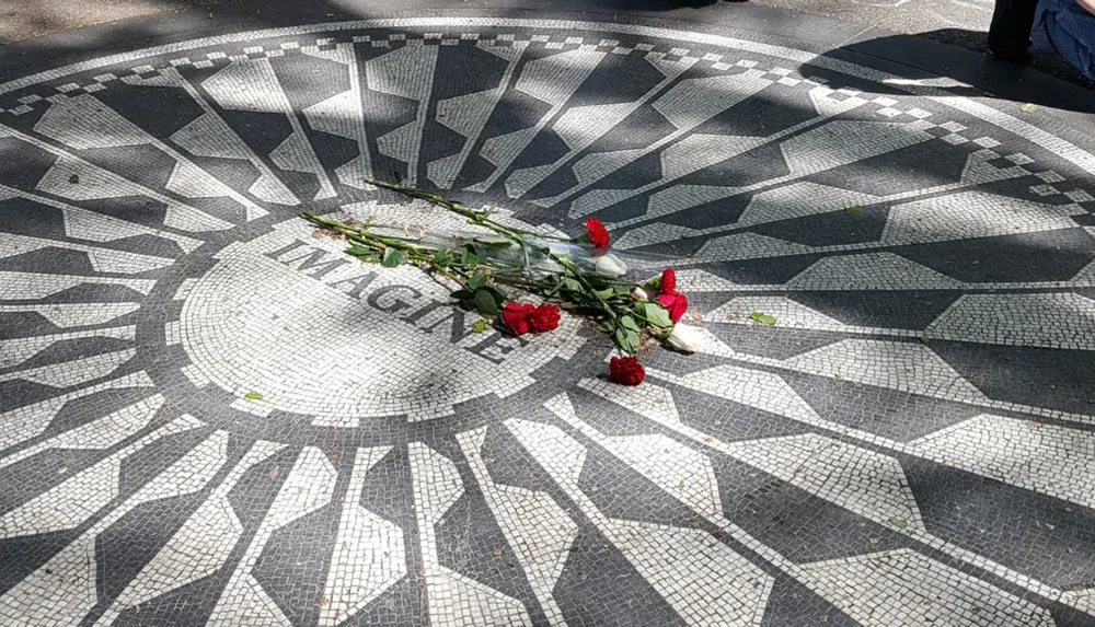 The image depicts a mosaic artwork with the word IMAGINE in the center adorned with a few scattered red roses likely serving as a memorial or tribute