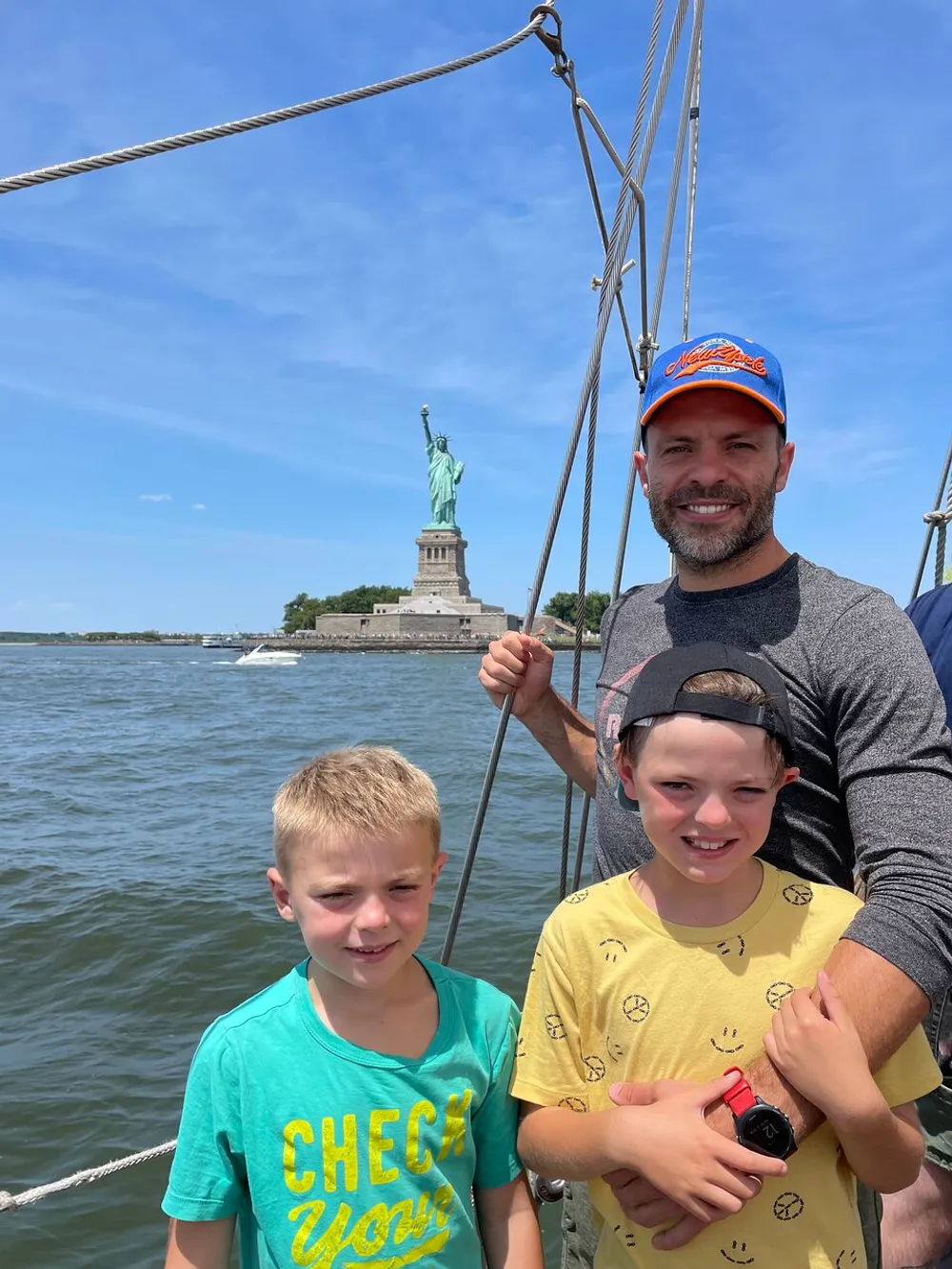 Three individuals are smiling on a sailboat with the Statue of Liberty in the background