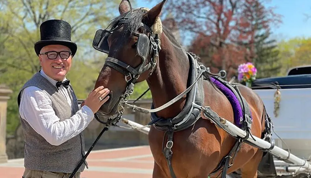 A smiling man in a top hat and traditional driving attire is standing next to and affectionately touching a well-groomed carriage horse