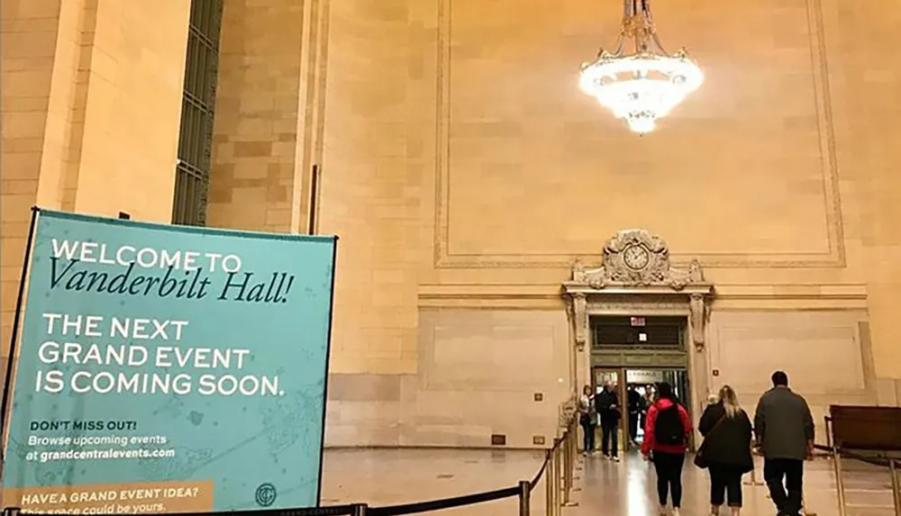 A promotional banner welcomes visitors to Vanderbilt Hall while people walk through a grand spacious interior lit by an elegant chandelier