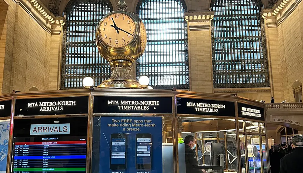 The image features the iconic golden clock in a grand train station hall with high arched windows and information booths displaying train arrivals and schedules