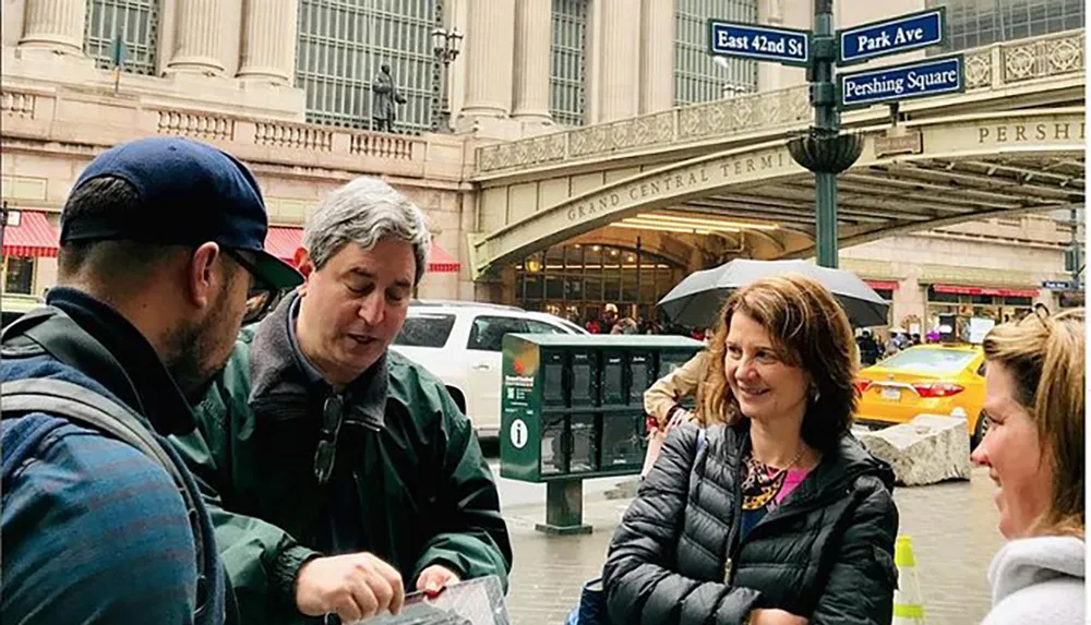 A group of people appears engaged in conversation near the entrance of Grand Central Terminal on a cloudy day with street signs for East 42nd Street and Park Avenue visible above them