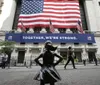A statue of a girl stands defiantly in front of the New York Stock Exchange which is adorned with an American flag and a banner saying TOGETHER WERE STRONG