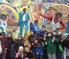 A group of smiling children poses in front of a colorful mosaic wall featuring artistic depictions of people