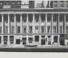 The image shows a historic black and white photograph of a city street with the facades of several buildings one being the Olympic Theatre surrounded by other businesses including a banner painter and a Masonic publishing company