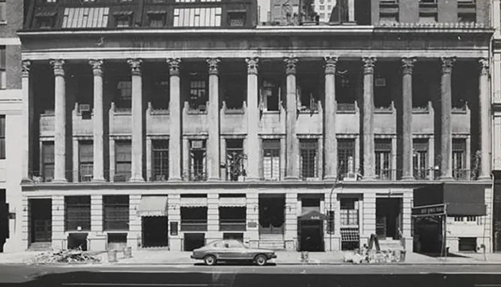 The image shows a black and white photograph of an older building with classical columns several boarded-up windows and a vintage car parked in front