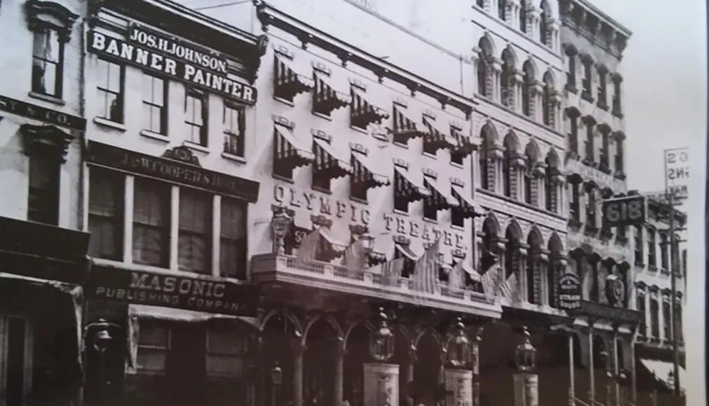 The image is a black and white photo of an old multi-story building with various business signs including Jos H Johnson Banner Painter and Olympic Theatre indicating a bustling commercial area from an earlier era