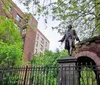 The image shows a statue of a historical figure atop a pedestal behind an ornate fence with leafy green trees and brick buildings in the background