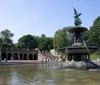 The image shows a tranquil scene at a large fountain with a statue on top surrounded by water and set against a backdrop of steps and archways with people leisurely enjoying the serene environment