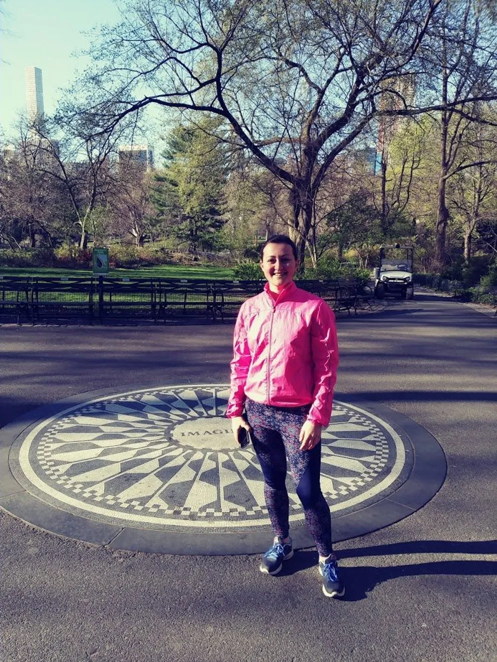 A person is standing on a circular patterned pavement with the word Imagine inscribed on it in a sunny park setting with trees and a high-rise building in the background
