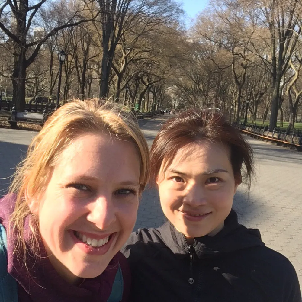 Two people are taking a smiling selfie on a sunny day on a tree-lined path likely in a park