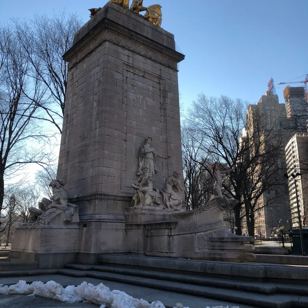 The image showcases a stone monument adorned with sculptural figures likely a historical or commemorative site set against a backdrop of bare trees and high-rise buildings with remnants of snow on the ground
