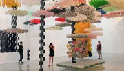 The image shows three people observing a large, intricate hanging artwork composed of various colorful patterned pieces in an exhibition space.