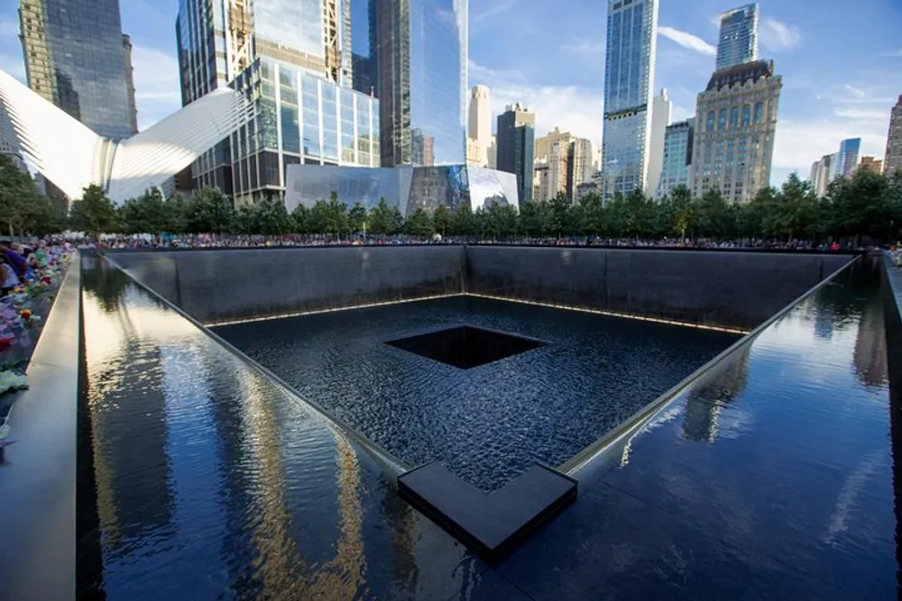 The image shows the 911 Memorial with one of its large reflective pools and the names of victims inscribed along the pools edges set against a backdrop of the New York City skyline