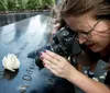 A woman is photographing a white rose placed on a name at a memorial with engraved names capturing a poignant moment of remembrance