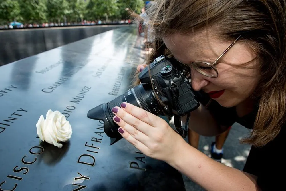 A woman is photographing a white rose placed on a name at a memorial with engraved names capturing a poignant moment of remembrance