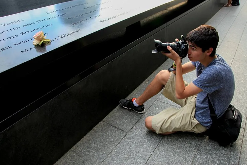 A person is crouching with a camera preparing to take a photograph of a single rose placed on a memorial with names engraved on it