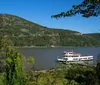 A sightseeing boat with passengers on deck is cruising on a river with a backdrop of a hillside displaying fall foliage