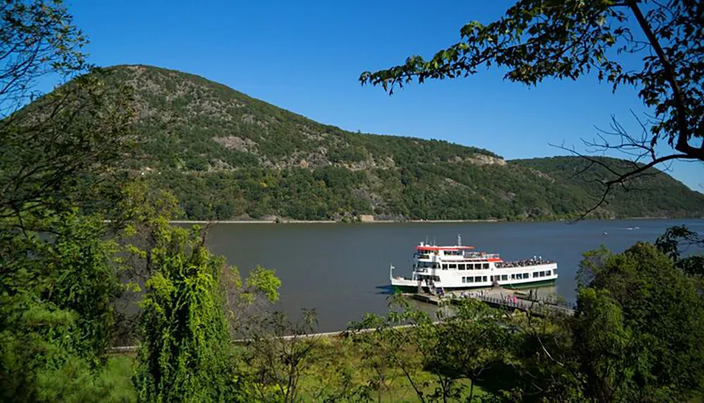 A sightseeing boat is docked by a river with lush green hills in the background on a clear day