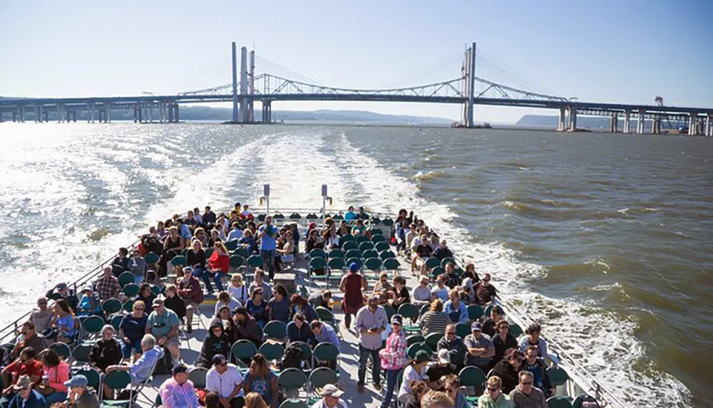 Passengers enjoy a sunny day on a ferry with a view of a large bridge in the background