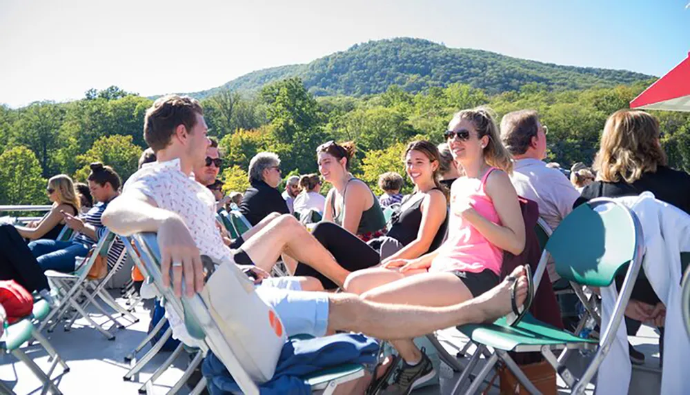 A group of people is enjoying a sunny day outdoors sitting on folding chairs and engaging in conversation with a backdrop of lush green hills