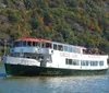 A sightseeing boat with passengers on deck is cruising on a river with a backdrop of a hillside displaying fall foliage