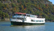 A sightseeing boat with passengers on deck is cruising on a river with a backdrop of a hillside displaying fall foliage.