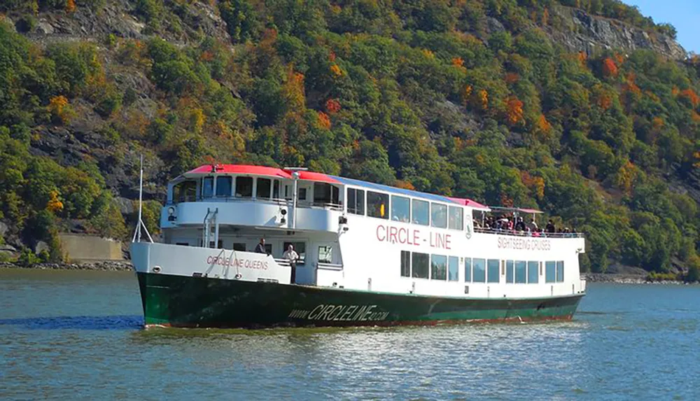 A sightseeing boat with passengers on deck is cruising on a river with a backdrop of a hillside displaying fall foliage