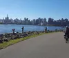 People are enjoying various outdoor activities along a riverside path with a city skyline in the background