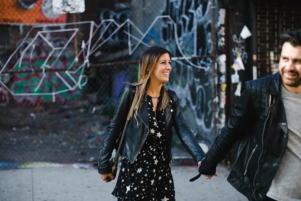 A woman in a star-patterned dress and a leather jacket is smiling while walking and holding hands with a man against a backdrop of graffiti-covered walls