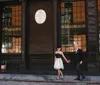 A well-dressed couple is holding hands and smiling at each other on a city sidewalk in front of a building with large windows and a classic architectural relief