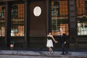 A well-dressed couple is holding hands and smiling at each other on a city sidewalk in front of a building with large windows and a classic architectural relief.