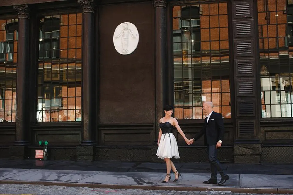 A well-dressed couple is holding hands and smiling at each other on a city sidewalk in front of a building with large windows and a classic architectural relief