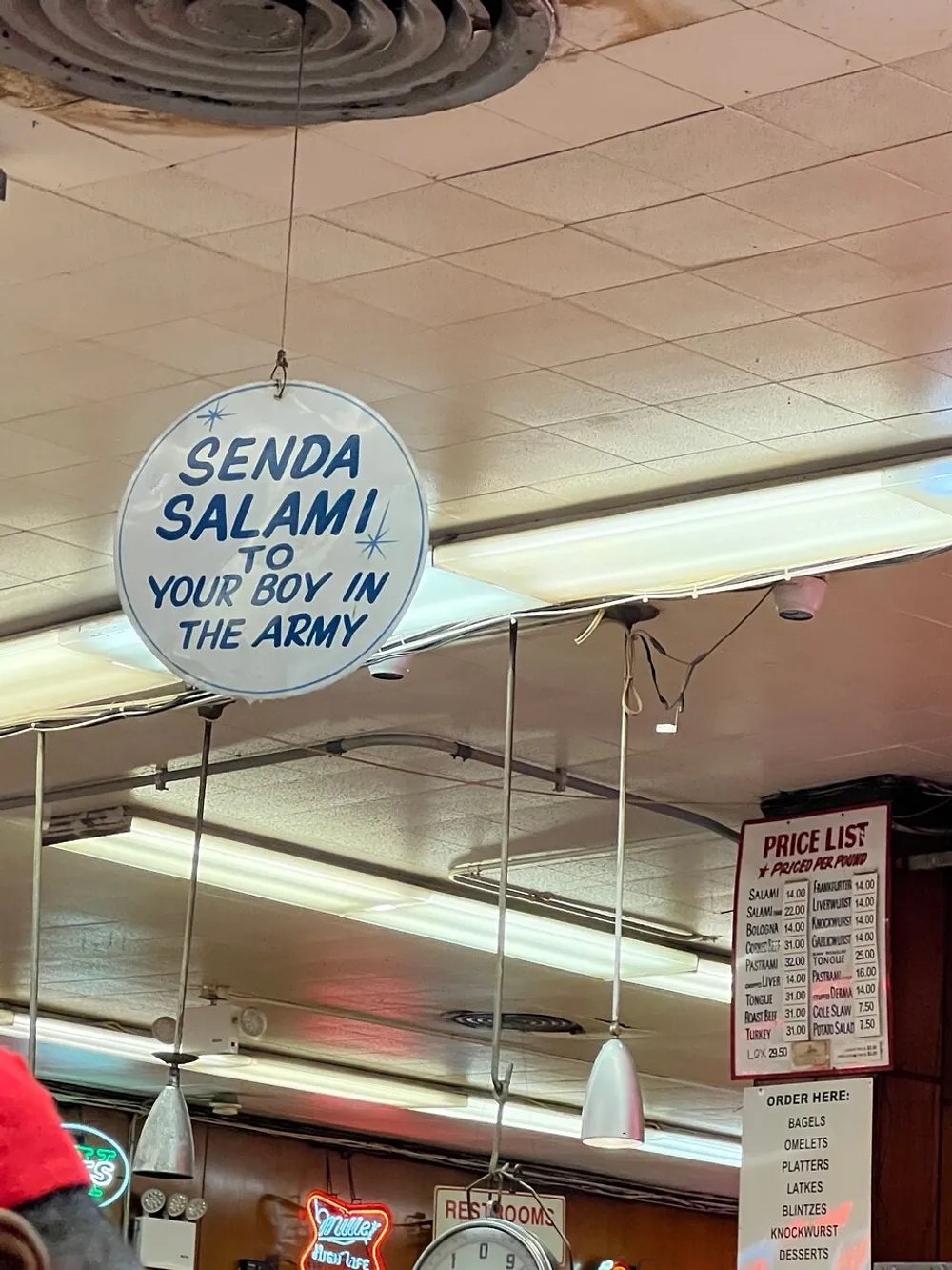 A sign hanging from the ceiling says SENDA SALAMI TO YOUR BOY IN THE ARMY suggesting a delis promotional offer in a nostalgic or supportive gesture for military service members