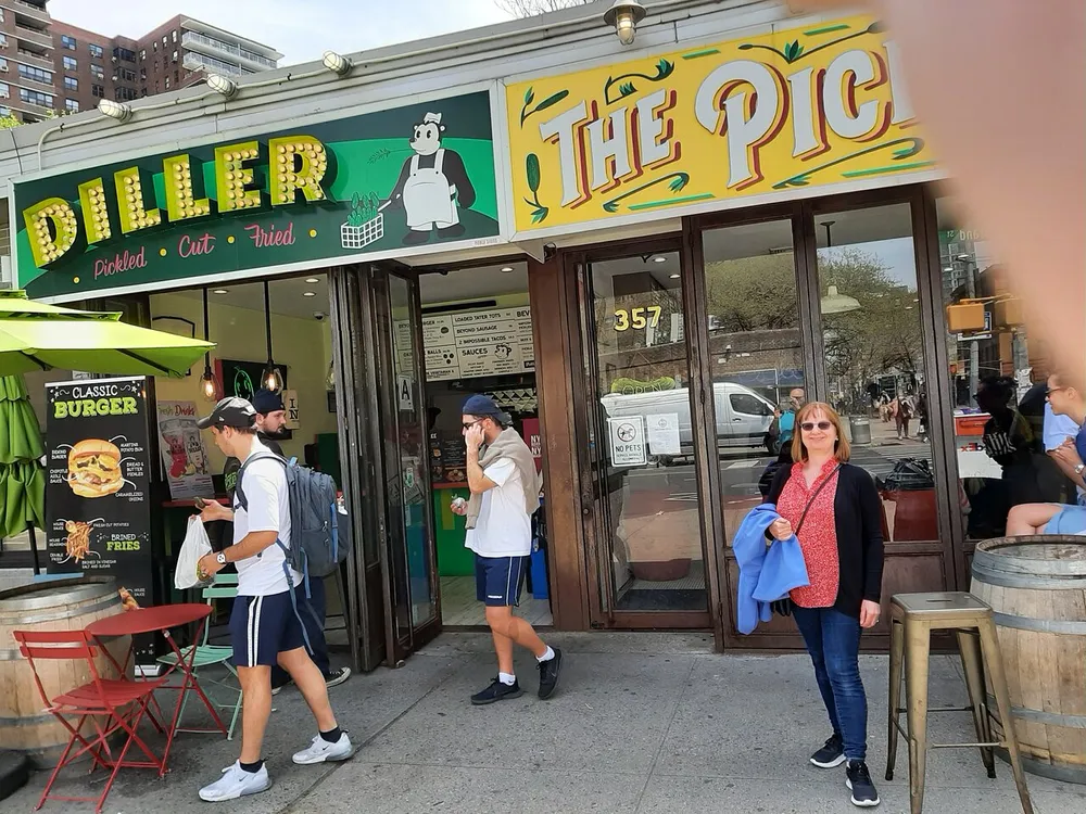 A woman stands in front of a bustling food establishment named The Pickle as patrons pass by capturing a lively urban dining atmosphere