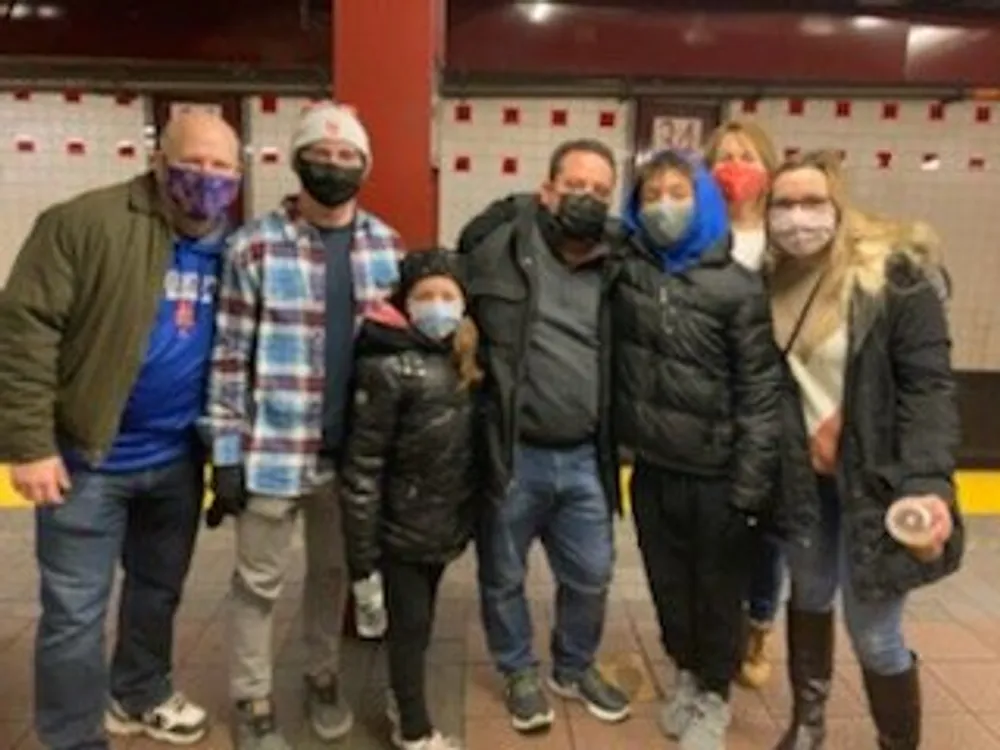 A group of seven individuals is posing for a photo in a subway station with several wearing face masks