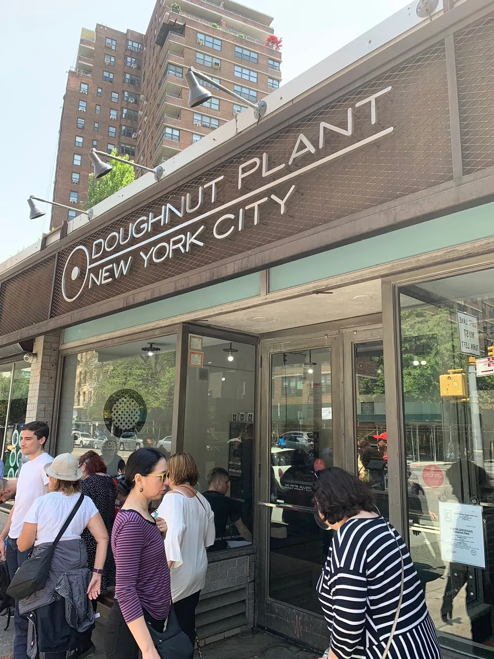 This image shows a group of people waiting outside the Doughnut Plant shop in New York City on a sunny day