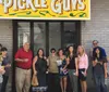 A group of people is standing in front of a store named The Pickle Guys holding pickles and smiling