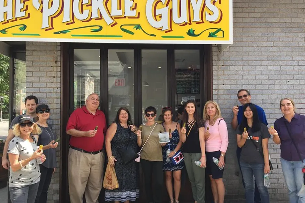 A group of people is standing in front of a store named The Pickle Guys holding pickles and smiling